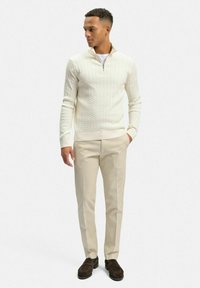 Krämfärgad kabelstickad tröja med halvdragkedja, parat med ljusbeiga byxor och mörkbruna loafers, stående mot en neutral bakgrund.