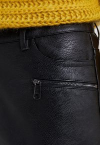 Czarne skórzane spodnie o teksturowanym wykończeniu, z kieszenią na zamek błyskawiczny i kontrastującym, żółtym swetrem dzianinowym na górze.