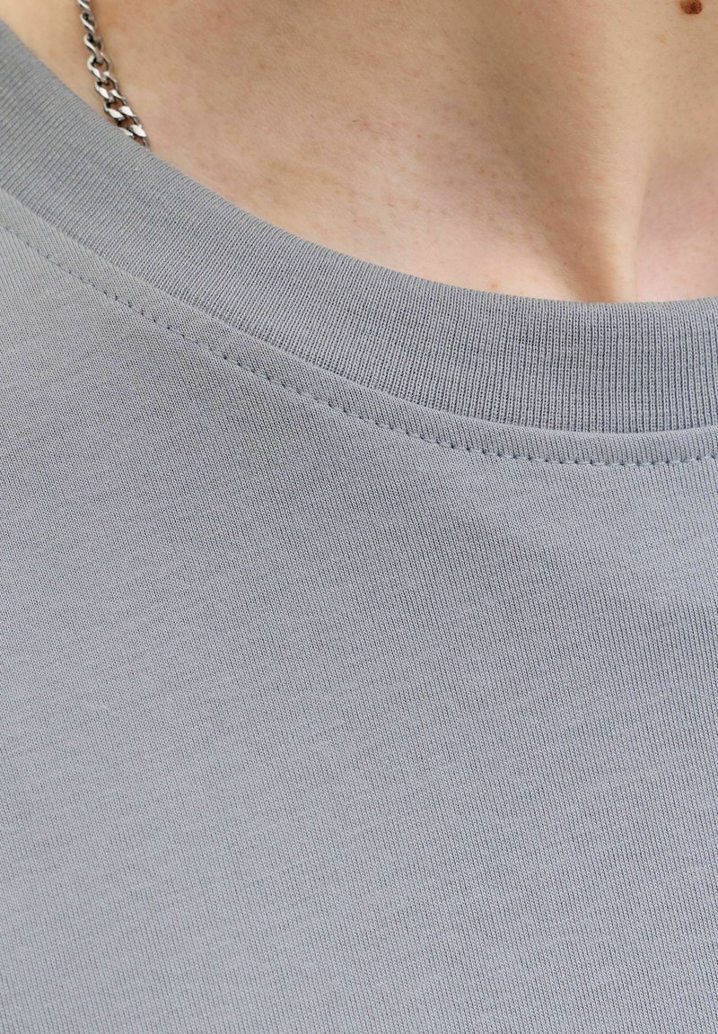 Közeli kép egy személy nyakáról, aki szürke kereknyakú pólót visel, valamint egy ezüst lánc nyakláncot, világos bőrszínnel és egy jól látható anyajeggyel.