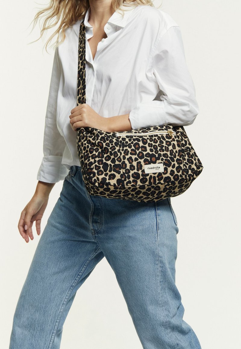 Femme portant une chemise blanche et un jean bleu, tenant un sac à épaule en imprimé léopard avec une poche zippée à l'avant et un patch logo.