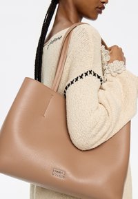 Tan leather tote bag with smooth texture and a silver logo accent. The model wears a cream sweater with black embroidery.
