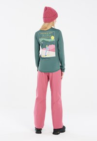 Langarm groene top met skisportafbeelding op de achterkant, gecombineerd met roze loszittende broek en zwarte laarzen. Gezellige gebreide roze muts.