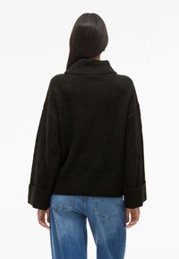 Suéter oversized verde escuro com um colarinho alto, punhos canelados e textura suave; combinado com jeans de denim azul, visto de trás.