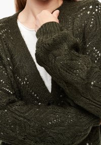 Olivgrüner, gestrickter Cardigan mit strukturiertem Muster, V-Ausschnitt und oversized Ärmeln. Ein schwarzer Knopf sichtbar.