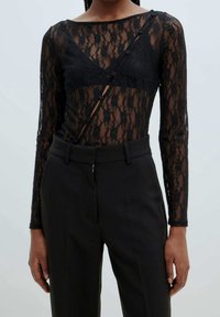 Haut à manches longues en dentelle noire avec un col en V, présentant une texture transparente et des boutons décoratifs sur un côté. Associé à un pantalon noir ajusté.