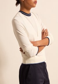 Osoba z założonymi rękami, ubrana w kremowy kardigan z granatowym i jasnoniebieskim wykończeniem, pod którym nosi dopasowaną bluzkę i ciemnogranatowe spodnie.