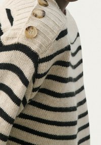 Kermanvärinen villapaita mustilla raidoilla, neulottu rakenne, resorikaulus ja kolme koristeellista nappia olkapäällä.