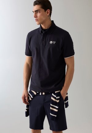 Młody mężczyzna w granatowej koszulce polo i szortach z paskowanym swetrem wiązanym wokół talii, stojący na tle jednolitego jasnego tła.