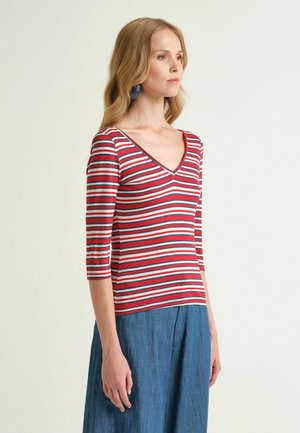 Donna con capelli biondi che indossa orecchini blu, una maglietta a righe rosse, bianche e blu navy con scollo a V e maniche a tre quarti, e pantaloni blu.