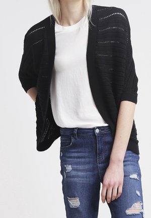 Personne portant un t-shirt blanc, un cardigan noir à maille ouverte et un jean bleu déchiré, debout avec une main derrière le dos.