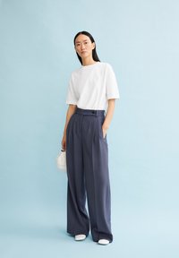 Vit t-shirt med kort ärm och liten logga, ihop med högmidjade, marinblå vida byxor. Modell som håller i en vit väska, stående mot en blå bakgrund.