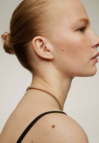 Vista de perfil de una persona con cabello corto y castaño claro recogido en un moño, vistiendo una blusa sin mangas negra y un collar de cadena dorada.