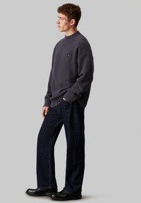 Maglione in microfibra grigio scuro con collo rotondo e orlo a coste, abbinato a jeans a gamba larga blu scuro e scarpe in pelle nere.