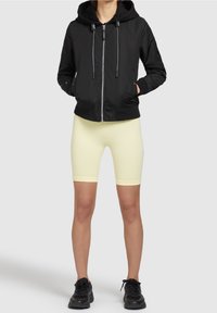 Czarna kurtka zapinana na zamek z kapturem, połączona z jasnobezowymi szortami rowerowymi i czarnymi butami sportowymi. Gładki materiał z kontrastującymi elementami.