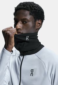 Black neck gaiter worn by a model, made of soft fabric. The model wears a white long-sleeve top with a black zip and logo.