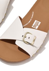 Witte slide-sandalen met een brede band, gouden gespdetail en een gevoerde bruine binnenzool. Bevat een gestructureerde voetbed en ronde studs op de zool.