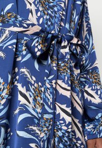 Blaues Kleid mit floralem Muster in Blau-, Weiß- und Orangetönen, mit einem gebundenen Stoffgürtel an der Taille und langen Ärmeln.