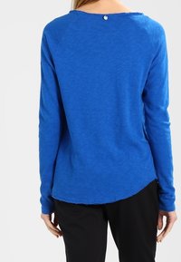 Top blu con maniche lunghe e scollatura arrotondata, caratterizzato da un piccolo bottone sul retro e un orlo leggermente curvo. Morbido al tatto.