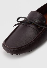 Mocasines de cuero marrón oscuro con un acabado texturizado, detalles cosidos y suela de goma negra con tachuelas. Presenta un lazo en la parte frontal y acentos de ojales.