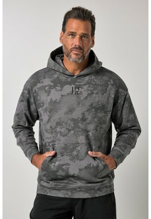 Homme avec barbe portant un sweat à capuche camouflage gris et noir avec un symbole mathématique imprimé, debout devant un fond uni.