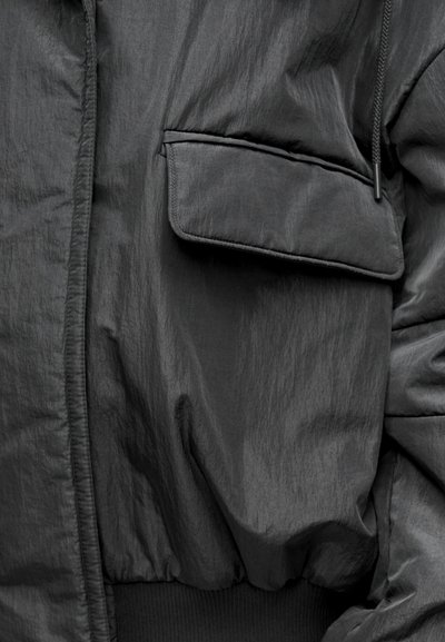 Juodas bombonešio tipo švarkas su tekstūruotu paviršiumi, turi šonines kišenes, elastinę apačią ir raištelį ant apykaklės.