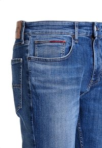 Siniset farkut, joissa on suora leikkaus, näkyvät ompeleet, nahkainen etiketti vyötäröllä sekä viisi taskua, joissa on punainen yksityiskohta.