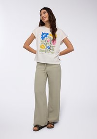 T-shirt a maniche corte color crema con stampa floreale in blu, rosa, giallo e verde. Abbinato a pantaloni larghi verde pallido e sandali neri.