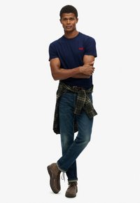 Man wearing a navy blue t-shirt, dark jeans, brown boots with laces, and a green plaid shirt tied around his waist, standing with arms crossed.