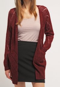 Burgundy stickad kappa med öppen fram, långa ärmar, diamantmönster och två framfickor, bärs över en ljus topp och svart kjol.