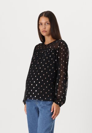 Blouse noire transparente avec des pois métalliques, décolleté rond et longues manches avec des poignets froncés, associée à un jean bleu clair.