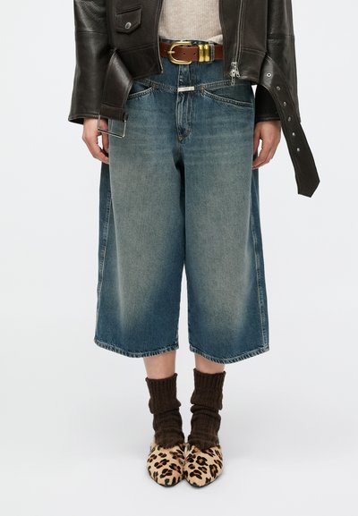 Culottes en denim bleu à jambes larges avec une finition délavée, associées à des chaussettes côtelées marron et des chaussures imprimé léopard. Stylisées avec une ceinture marron et une veste en cuir.