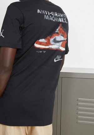 T-shirt in cotone nero con un'illustrazione colorata di una sneaker sul retro, con il testo "MACCHINE ANTIGRAVITÀ" e il marchio Nike.