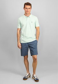 Ljust grön polo-shirt med krage, marinblå shorts och sneakers med vita snören och bruna detaljer. Enkel och avslappnad design.