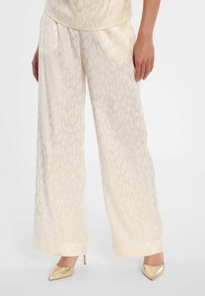 Vrouw draagt ivoorkleurige satijnen broek met wijde pijpen, subtiele luipaardprint en gouden puntige hoge hakken, staande tegen een effen achtergrond.