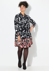 Abito nero floreale con fiori bianchi e dettagli rossi, maniche lunghe e gonna a campana. Abbinato a collant neri e scarpe.