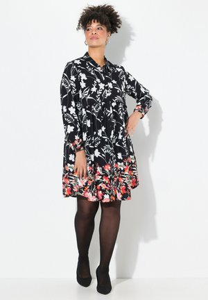 Schwarzes Blumenkleid mit weißen Blumen und roten Akzenten, langen Ärmeln und einem schwingenden Rock. Kombiniert mit schwarzen Strumpfhosen und Schuhen.