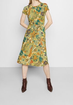 Femme portant une robe fleurie mi-longue à manches courtes avec des motifs jaunes, verts et orange, associée à des bottines marron, debout les mains derrière le dos.