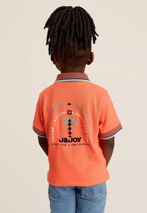 Kind mit lockigem Haar, das ein oranges Poloshirt mit Palmenmotiv und Schriftzug trägt, kombiniert mit blauen Jeans, mit dem Rücken zur Kamera.