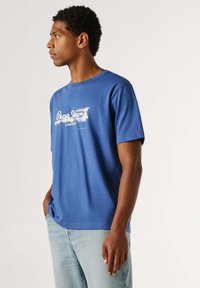 T-shirt en coton bleu avec col rond et manches courtes, affichant un logo blanc sur la poitrine et une coupe décontractée.