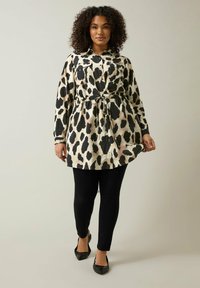 Tiermuster Kleid in Beige und Schwarz, in der Taille mit einem Bindegürtel gerafft. Lange Ärmel, zwei frontale Taschen und ein geschwungener Saum. Tragen mit schwarzen Leggings.