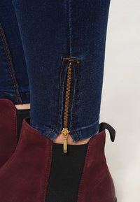 Vaqueros azules con un zipper dorado en el tobillo, combinados con botines de ante burdeos que tienen paneles laterales de elastano negro.
