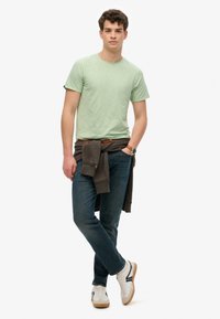 Camiseta verde de manga corta con un acabado texturizado, combinada con unos jeans azul oscuro. Un suéter marrón está atado a la cintura, complementado con zapatillas blancas.
