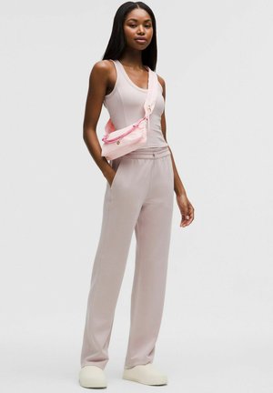 Ženska stoji v bledo roza zgornji majici brez rokavov in ujemajočih širokih hlačah z roza torbico čez ramo in belimi čevlji na zaponke.