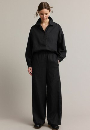 Vrouw met bruin haar in een zwart, knoopjeshemd met lange mouwen en wijde broekspijpen, staand met handen in de zakken tegen een effen achtergrond.