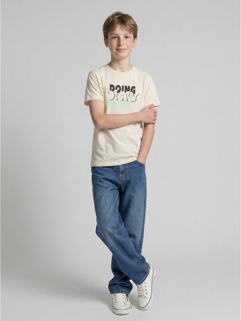 Dreng stående med krydsede ben iført en creme t-shirt med teksten "DOING", blå jeans og hvide sneakers på en ensfarvet grå baggrund.