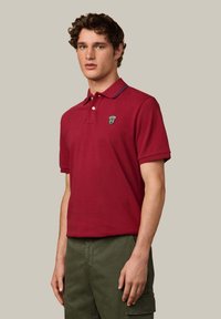 Červené polo tričko s golierom a dvomi gombíkmi na zapínanie. Obsahuje logo na hrudi a kontrastné námornícke akcenty na golieri a rukávoch.