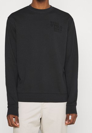 Mann trägt schwarzen Marc O’Polo Denim Sweatshirt mit langen Ärmeln und beige Hose, steht vor einem schlichten hellen Hintergrund.