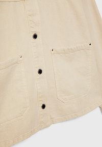Vêtement beige avec deux poches avant et des boutons-pression noirs au centre, en tissu texturé.