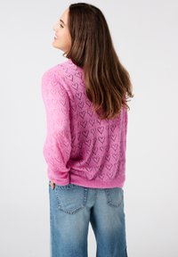 Roze opengewerkt sweater met een hartpatroon, lange mouwen en geribde manchetten. Gecombineerd met lichtblauwe wijde jeans.