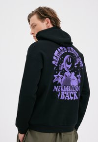Moški, oblečen v črno kapuco s purpurnim grafikonom nasmejanega lika, zvezd in besedilom "ALWAYS KEEP ON" in "NEVER LOOK BACK" na hrbtu.